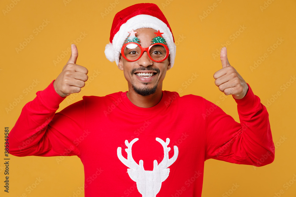Smiling young Santa african american man 20s in red sweater Christmas hat glasses showing thumbs up isolated on yellow background studio portrait. Happy New Year celebration merry holiday concept. Smiling young Santa african american man 20s in red sweater Christmas hat glasses showing thumbs up isolated on yellow background studio portrait. Happy New Year celebration merry holiday concept.