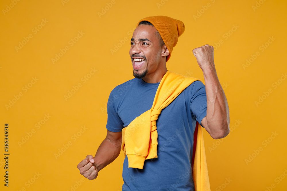 Overjoyed young african american man 20s in basic casual blue t-shirt hat standing clenching fists doing winner gesture looking aside isolated on bright yellow colour background, studio portrait.