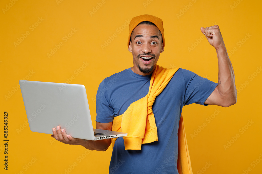 Excited joyful happy young african american man 20s wearing casual blue t-shirt hat standing using laptop pc computer doing winner gesture isolated on bright yellow colour background, studio portrait. Excited joyful happy young african american man 20s wearing casual blue t-shirt hat standing using laptop pc computer doing winner gesture isolated on bright yellow colour background, studio portrait.