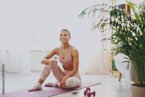 Smiling beautiful attractive young strong sporty fitness woman wearing pink tracksuit doing yoga exercises sitting holding bottle of water stretching on mat floor at home indoor.