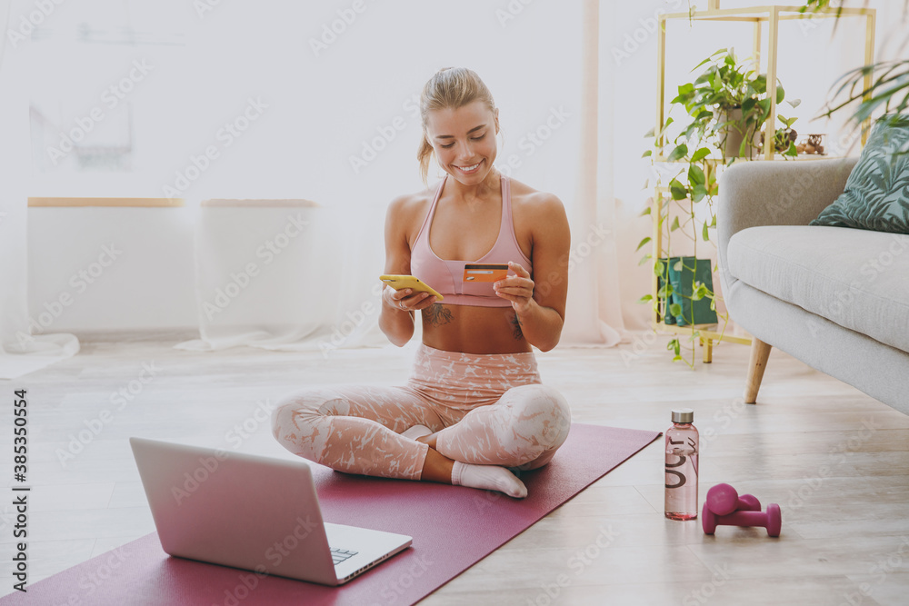 Smiling young strong sporty fitness woman wearing pink tracksuit doing yoga exercises using laptop mobile phone hold credit bank card showing thumb up sitting stretching on mat floor at home indoor. Smiling young strong sporty fitness woman wearing pink tracksuit doing yoga exercises using laptop mobile phone hold credit bank card showing thumb up sitting stretching on mat floor at home indoor.