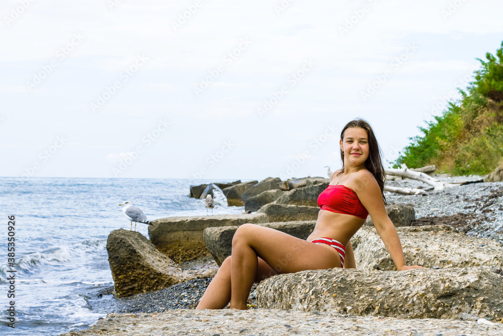 A young girl in a red swimsuit sits on the stones on the seashore with seagulls.