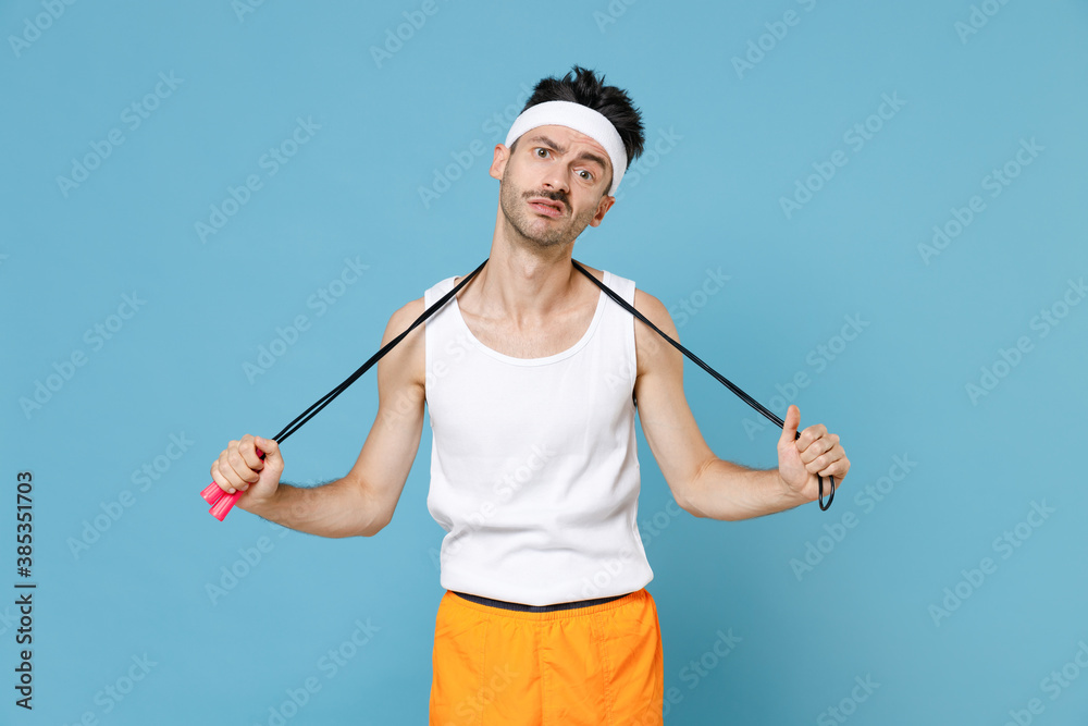 Exhausted confused young sporty man with thin skinny body sportsman in headband shirt shorts standing with skipping rope over neck isolated on blue background. Workout gym sport motivation concept. Exhausted confused young sporty man with thin skinny body sportsman in headband shirt shorts standing with skipping rope over neck isolated on blue background. Workout gym sport motivation concept.