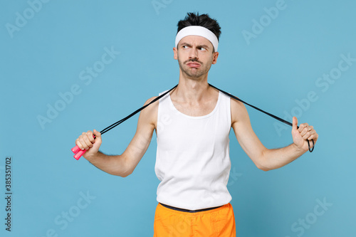 Tired confused young sporty man with thin skinny body sportsman in white headband shirt shorts standing with skipping rope over neck isolated on blue background. Workout gym sport motivation concept.