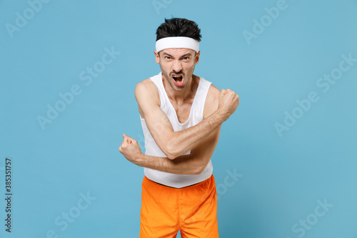 Screaming strong young sporty fitness man with thin skinny body sportsman in white headband shirt shorts showing biceps muscles isolated on blue background. Workout gym sport motivation concept.