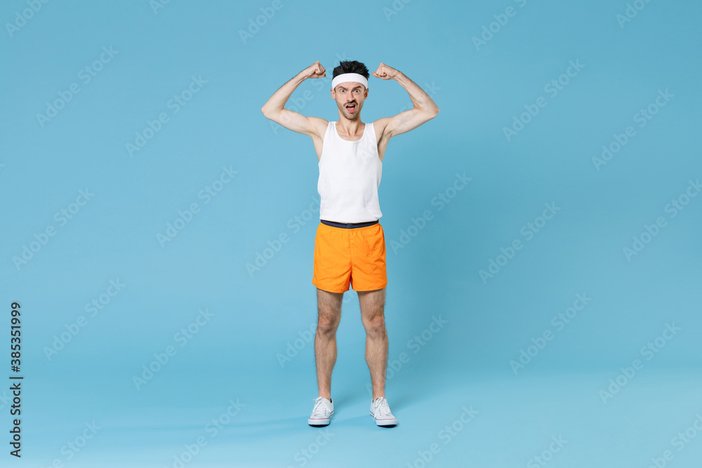 Full length portrait of shocked strong young fitness man with skinny body sportsman in headband shirt shorts showing biceps muscles isolated on blue background. Workout gym sport motivation concept.