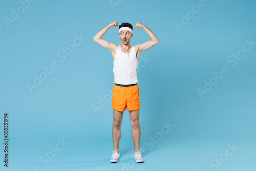 Full length portrait of amazed strong young fitness man with skinny body sportsman in headband shirt shorts showing biceps muscles isolated on blue background. Workout gym sport motivation concept.