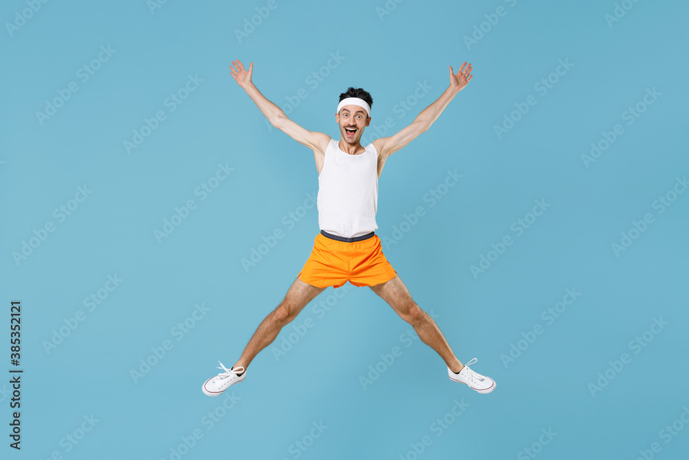 Full length portrait surprised young fitness man with skinny body sportsman in headband shirt shorts jumping spreading hands and legs isolated on blue background. Workout gym sport motivation concept.