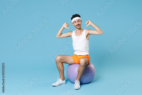 Full length portrait excited young man with skinny body sportsman in headband shirt shorts sit on fitball pointing thumbs on himself isolated on blue background. Workout gym sport motivation concept.