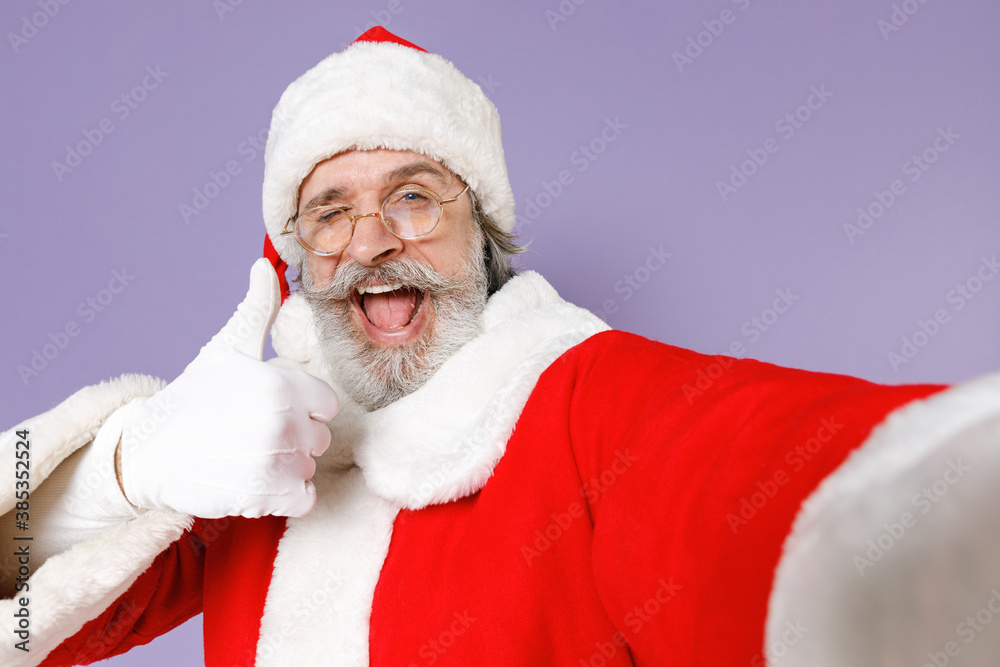 Naklejka premium Close up blinking Santa Claus man in Christmas hat red suit glasses doing selfie shot on mobile phone showing thumb up isolated on violet background. Happy New Year celebration merry holiday concept.