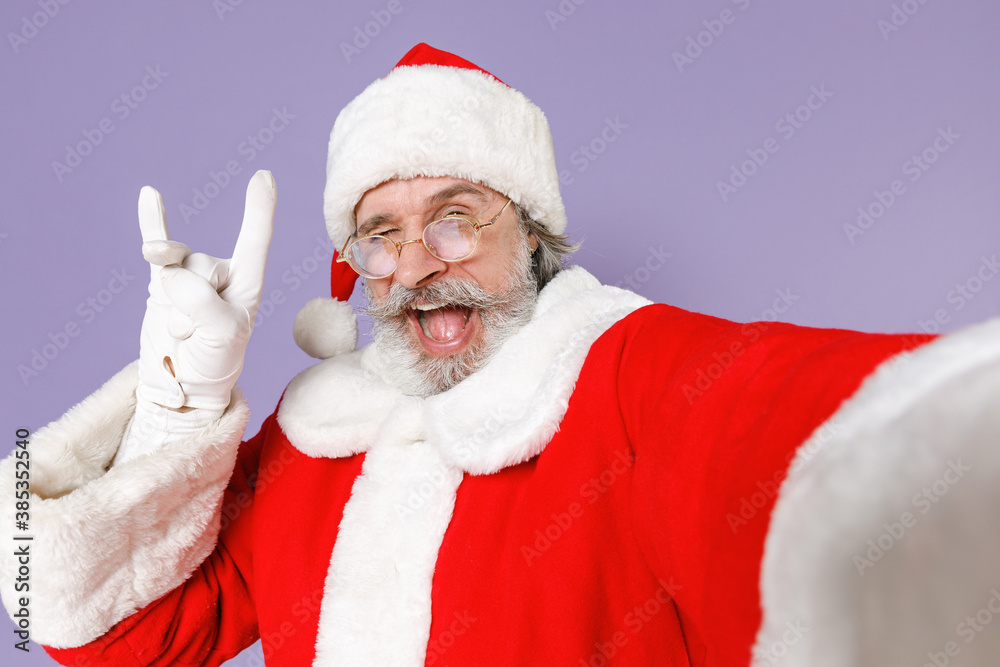 Naklejka premium Close up of funny Santa Claus man in Christmas hat red suit doing selfie shot on mobile phone showing horns up gesture isolated on violet background. Happy New Year celebration merry holiday concept.