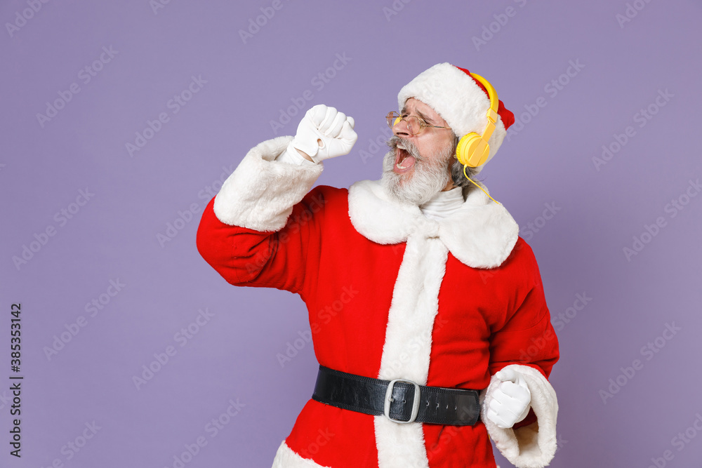 Cheerful Santa Claus man in Christmas hat red suit coat white gloves glasses  listening music with headphones sing song isolated on violet background.  Happy New Year celebration merry holiday concept. Photos