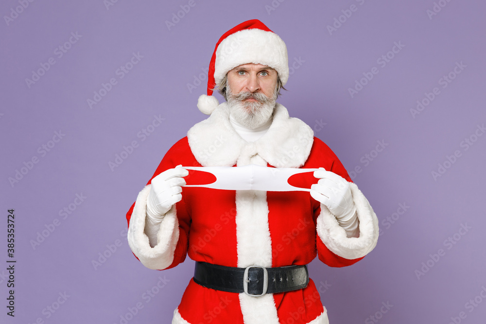 Displeased Santa Claus man in Christmas hat red coat hold face mask to safe from coronavirus virus covid-19 during quarantine isolated on violet background. New Year celebration merry holiday concept. Displeased Santa Claus man in Christmas hat red coat hold face mask to safe from coronavirus virus covid-19 during quarantine isolated on violet background. New Year celebration merry holiday concept.