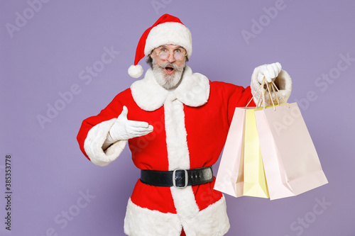 Shocked Santa Claus man in Christmas hat red coat suit glasses pointing hand on package bags purchases after shopping isolated on violet background. Happy New Year celebration merry holiday concept.