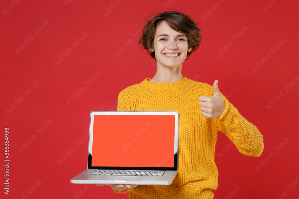 Smiling young brunette woman 20s wearing yellow sweater standing hold laptop pc computer with blank empty screen mock up copy space showing thumb up isolated on bright red background studio portrait. Smiling young brunette woman 20s wearing yellow sweater standing hold laptop pc computer with blank empty screen mock up copy space showing thumb up isolated on bright red background studio portrait.