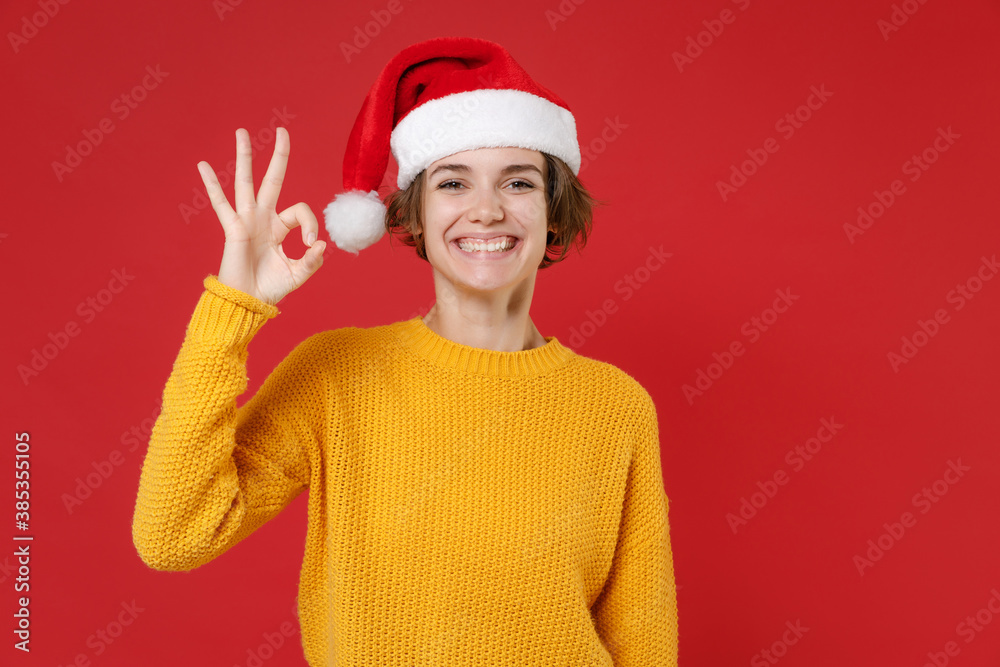 Smiling young Santa woman 20s wearing casual yellow sweater Christmas hat showing Ok gesture isolated on bright red colour background studio portrait. Happy New Year celebration merry holiday concept. Smiling young Santa woman 20s wearing casual yellow sweater Christmas hat showing Ok gesture isolated on bright red colour background studio portrait. Happy New Year celebration merry holiday concept.