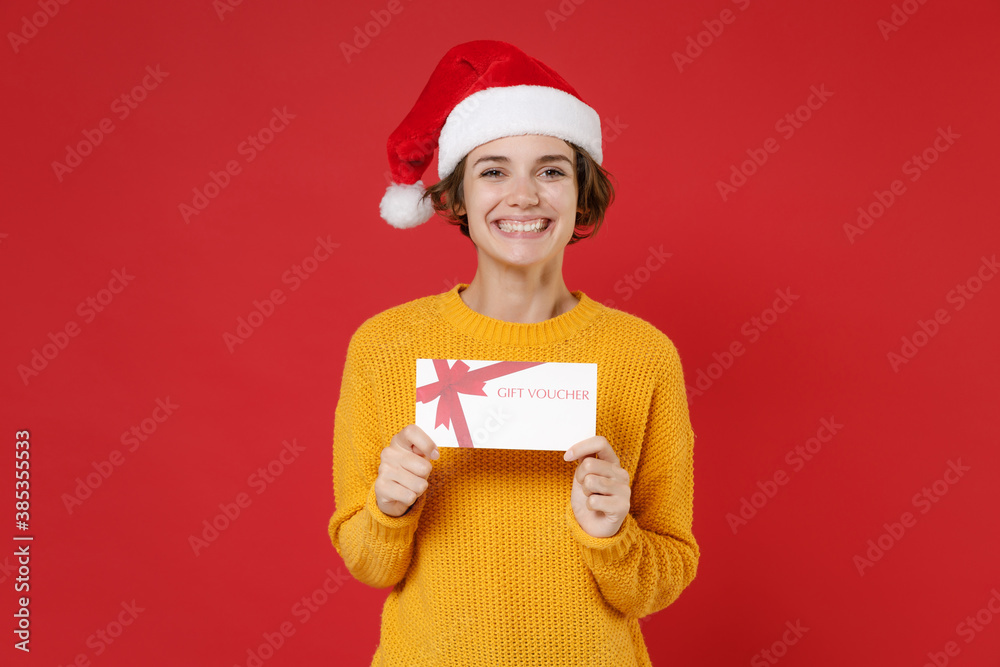 Smiling young Santa woman wearing casual yellow sweater Christmas hat hold gift certificate looking camera isolated on red background studio portrait. Happy New Year celebration merry holiday concept. Smiling young Santa woman wearing casual yellow sweater Christmas hat hold gift certificate looking camera isolated on red background studio portrait. Happy New Year celebration merry holiday concept.