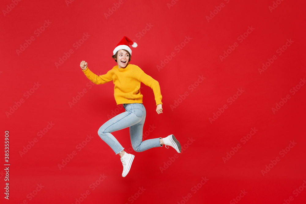 Full length side view of funny young Santa woman in yellow sweater Christmas hat jumping like running looking aside isolated on red background studio. Happy New Year celebration merry holiday concept.