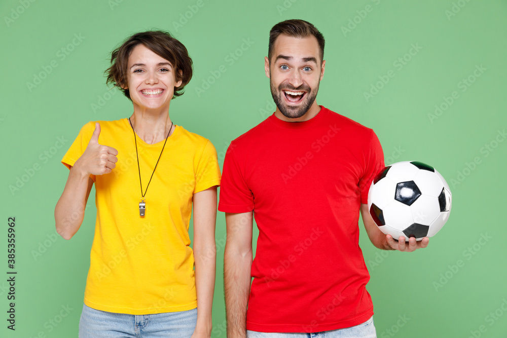 Smiling funny young couple friends sport family woman man football fans in yellow red t-shirts cheer up support favorite team with soccer ball showing thumb up isolated on green background studio. Smiling funny young couple friends sport family woman man football fans in yellow red t-shirts cheer up support favorite team with soccer ball showing thumb up isolated on green background studio.