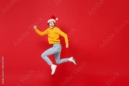 Full length side view of funny young Santa woman in yellow sweater Christmas hat jumping like running looking aside isolated on red background studio. Happy New Year celebration merry holiday concept.