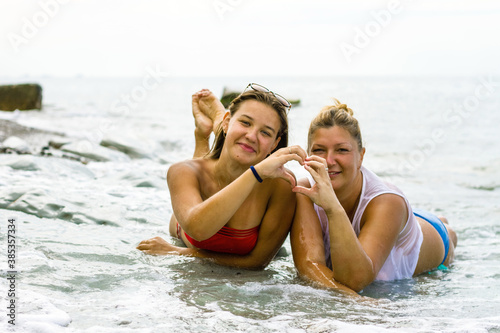 Two young women of different ages in bikinis lie on the seashore, the waves cover the girls © Vera