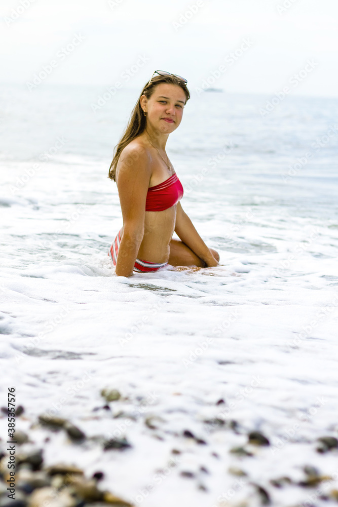 A young brunette girl in a red swimsuit sits on the seashore, the waves cover the knees and legs of the girl.