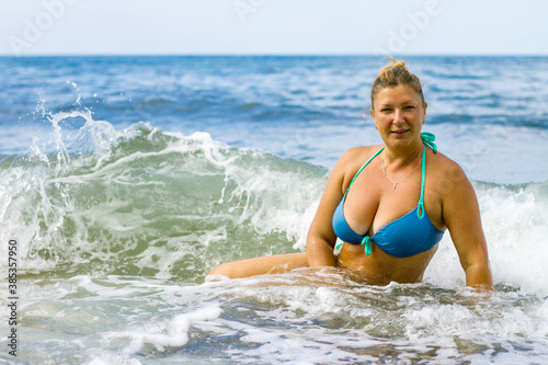 Young woman in a blue swimsuit sits on the seashore, waves and water cover the woman.