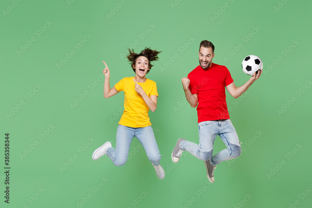 Full length of excited couple friends sport family woman man football fans cheer up support team with soccer ball jumping pointing index fingers up doing winner gesture isolated on green background.