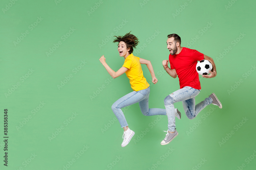Full length of excited young couple friends sport family woman man football fans in t-shirts cheer up support favorite team with soccer ball jumping like running isolated on green background studio.