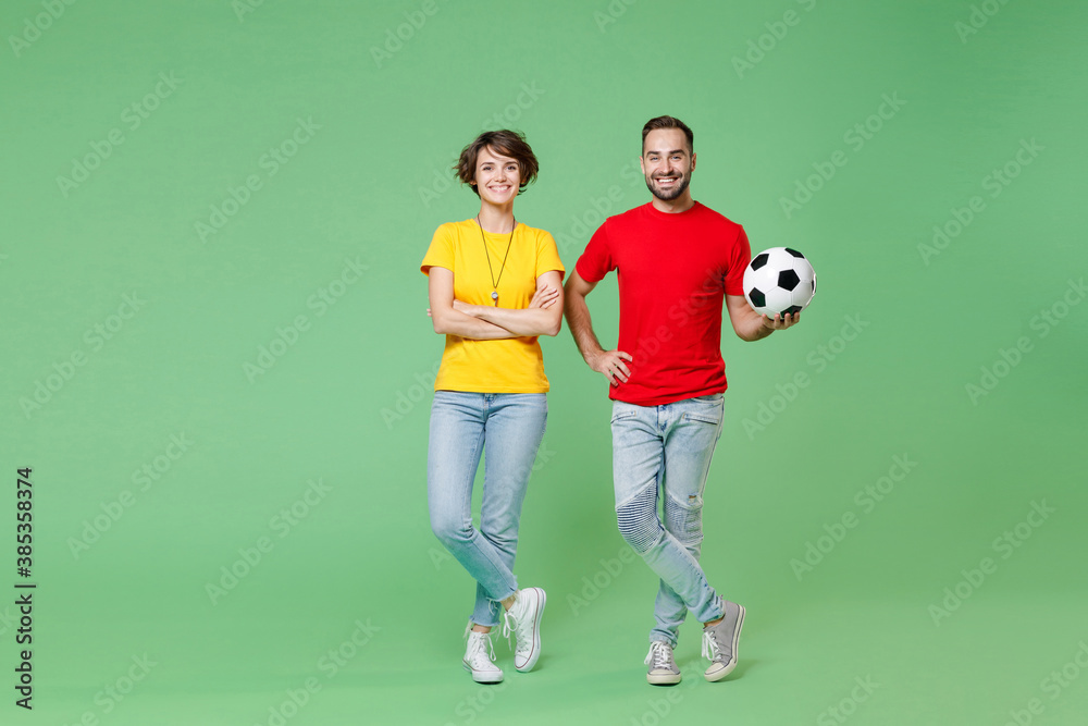 Full length portrait of smiling young couple friends sport family woman man football fans in yellow red t-shirts cheer up support favorite team with soccer ball isolated on green background studio.