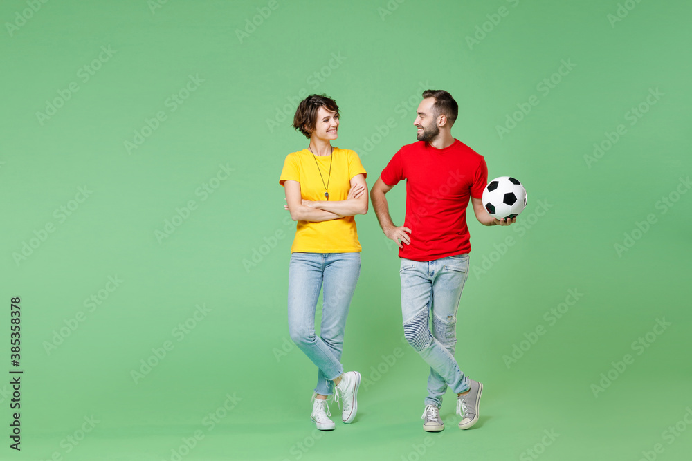 Full length smiling young couple friends sport family woman man football fans in yellow red t-shirts cheer up support favorite team with soccer ball looking at each other isolated on green background. Full length smiling young couple friends sport family woman man football fans in yellow red t-shirts cheer up support favorite team with soccer ball looking at each other isolated on green background.