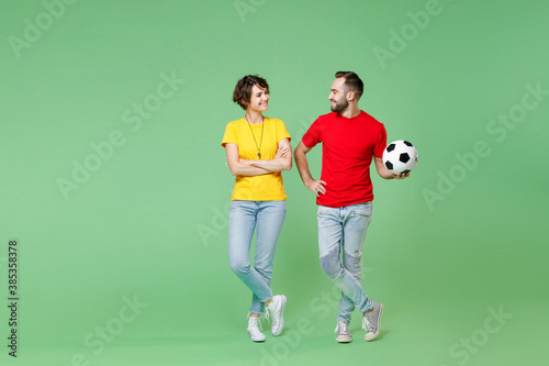 Fototapeta Naklejka Na Ścianę i Meble -  Full length smiling young couple friends sport family woman man football fans in yellow red t-shirts cheer up support favorite team with soccer ball looking at each other isolated on green background.