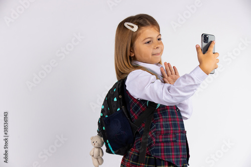Photo of joyful handsome blondine schoolgirl taking selfie on mobile phone and gesturing hello sign isolated on white background with free space