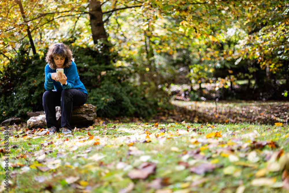 Autumn scene of middle aged woman wearing blue sweater sitting on stone and looking at smartphone next to pond covered in fallen leaves Autumn scene of middle aged woman wearing blue sweater sitting on stone and looking at smartphone next to pond covered in fallen leaves