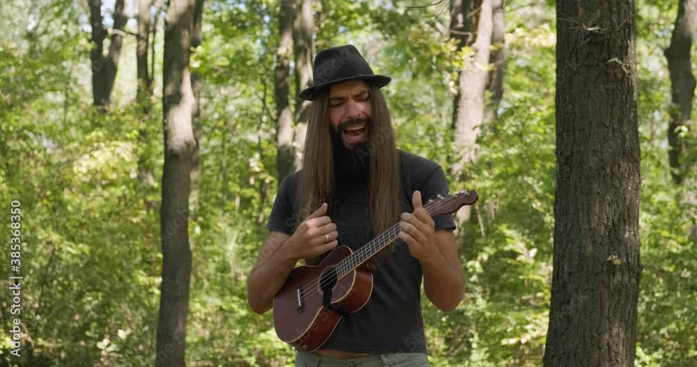 Bearded male with longhait and hat posing with ukulele in the forest Bearded male with longhait and hat posing with ukulele in the forest