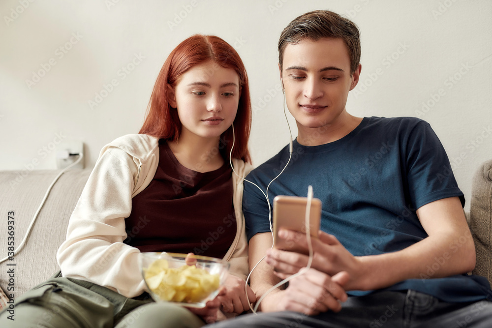 Couple of teenagers sitting on the couch at home, watching something on the phone, using the same pair of earphones while spending time together Couple of teenagers sitting on the couch at home, watching something on the phone, using the same pair of earphones while spending time together