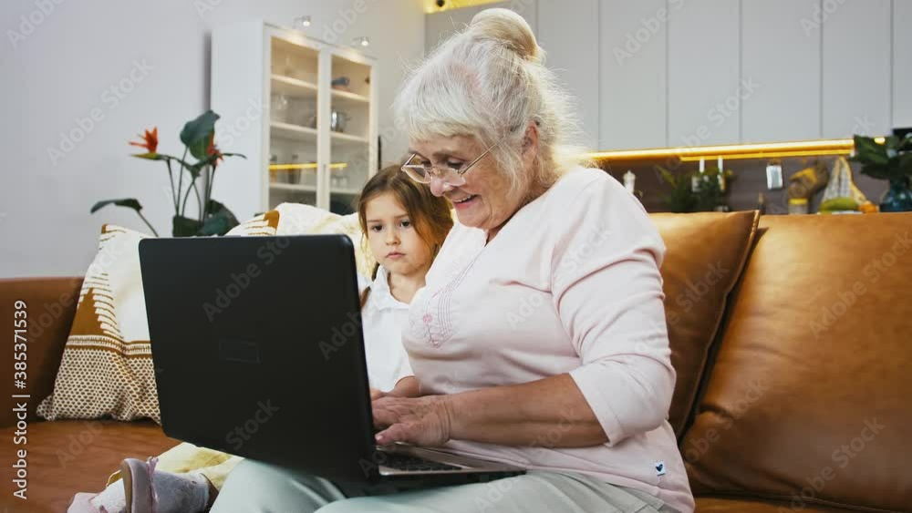 Grandma smiling, typing on laptop sitting with granddaughter on couch in studio flat with kitchen on background Grandma smiling, typing on laptop sitting with granddaughter on couch in studio flat with kitchen on background