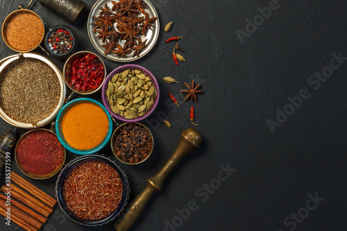 Bright spices and fragrant dried herbs in colored bowls, copper pestle for grinding herbs on a black background. Spices for making curry.Top view, with space. Concept of national cuisine. 