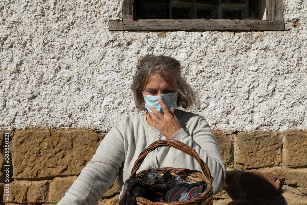 A senior woman, with short hair, sitting at the table in front of the mountain hut of the fountain of Artica, in Luesia, Spain, correctly adjusts her protective face mask.