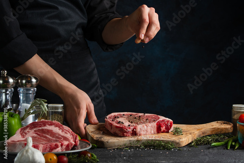 Close-up view of professional chef in black uniform salts raw steak on wooden chopped board. Backstage of preparing grilled pork meat at restaurant kitchen on dark blue background. Frozen motion.