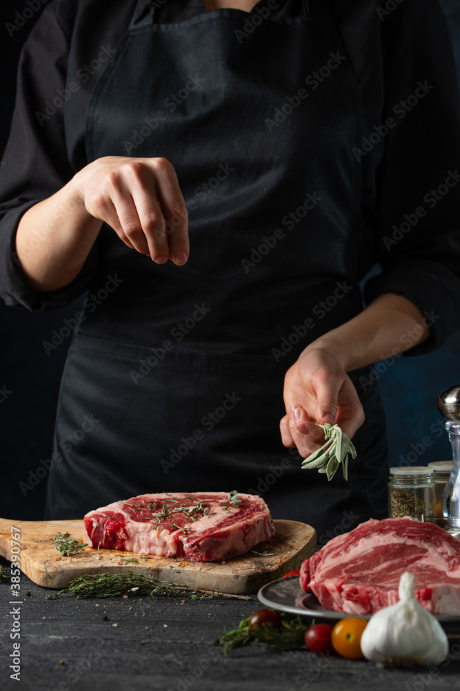 Close-up view of chef pours rosemary on raw steak on wooden chopped board. Backstage of preparing grilled pork meat at restaurant kitchen on dark blue background. Frozen motion. Vertical format.