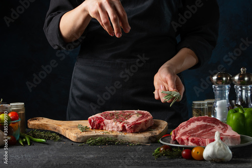 Close-up view of chef pours rosemary on raw steak on wooden chopped board. Backstage of preparing grilled pork meat at restaurant kitchen on dark blue background. Frozen motion. Horizontal format.