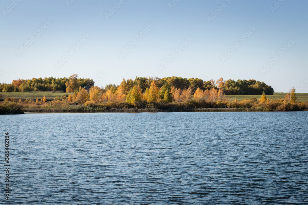 The blue sky is reflected in the waters of the lake. On the far shore there are yellow and green trees and bushes. Sunny autumn day.