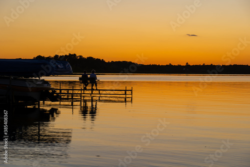 Evening scene at a lake in Bemidji, Minnesota, with silhouette of two people sitting on a dock bench, and a few ripples on the reflective water.