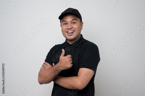 Smiling young handsome asian delivery man in black uniform standing with thumbs up on isolated white background.