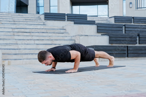 Man athlete in black sportswear practices yoga exercises in plank position outdoors.