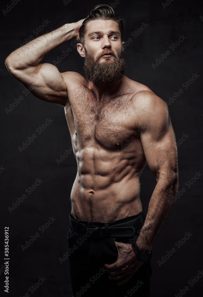 Brutal bearded guy with naked torso and muscular build with hand in pocket and hand under head posing in dark background.