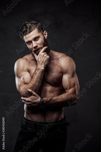Handsome and haired man with nude torso and muscular build posing with hand under his chin in dark background.