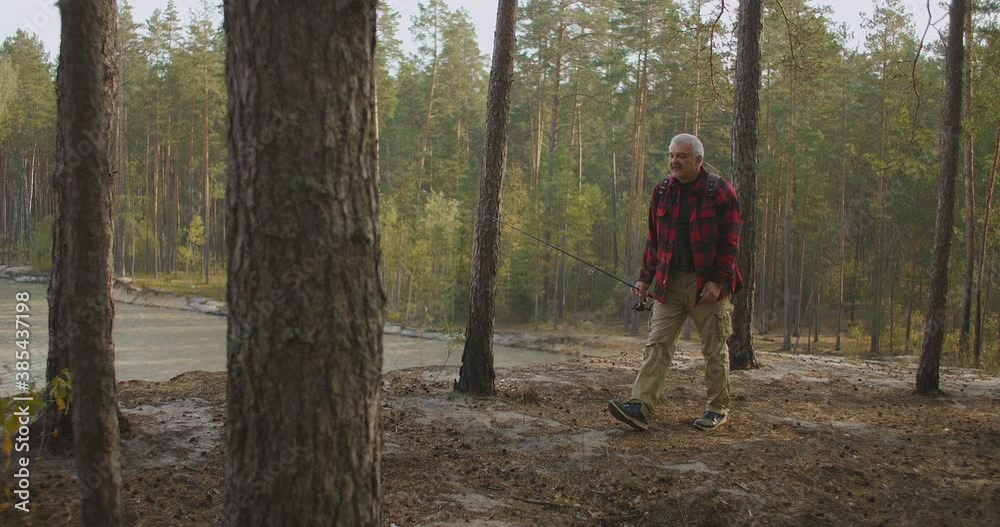 lonely aged hiker is walking in forest on high shore of reservoir, holding rod in hand, backpacking and angling lonely aged hiker is walking in forest on high shore of reservoir, holding rod in hand, backpacking and angling