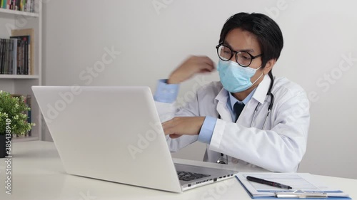 Young Asian Doctor Man in Lab Coat or Gown with Stethoscope Wear Face Mask and Glasses Exam Patient by Video Conference or Video Chat in Office